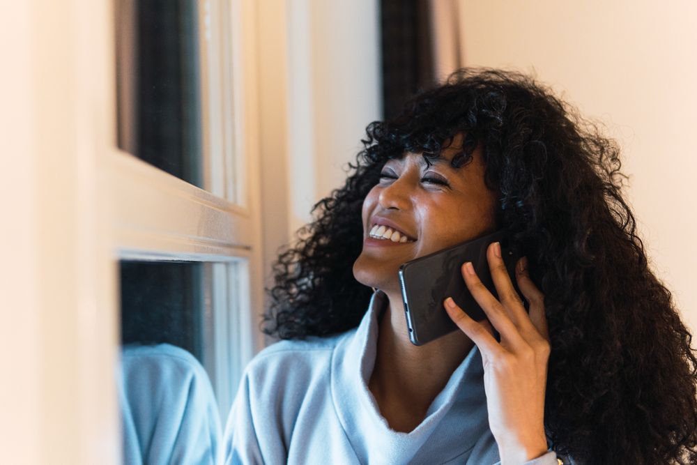 Cheerful Black woman talking smartphone at home