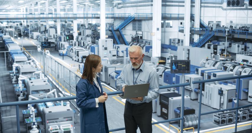 Male Engineer and Female Project Manager Standing on a Platform, Talking, Using Laptop Computer and Monitoring Production at a Modern Automated Electronics Manufacture with AI Robots