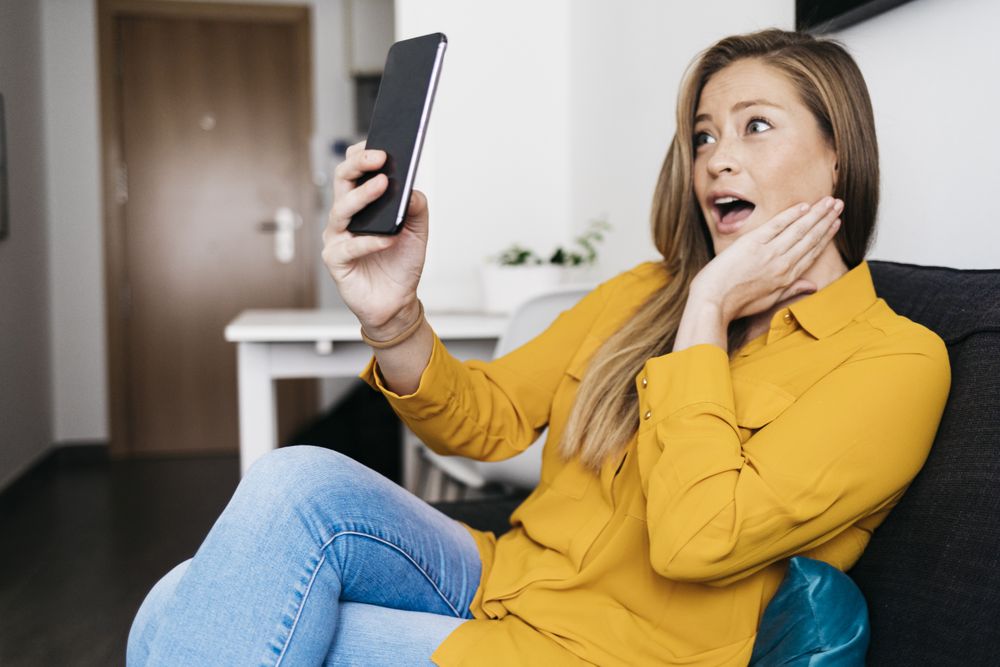 Surprised adult female sitting on sofa while using smartphone