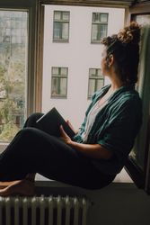 Interested woman reading book at window sill at home
