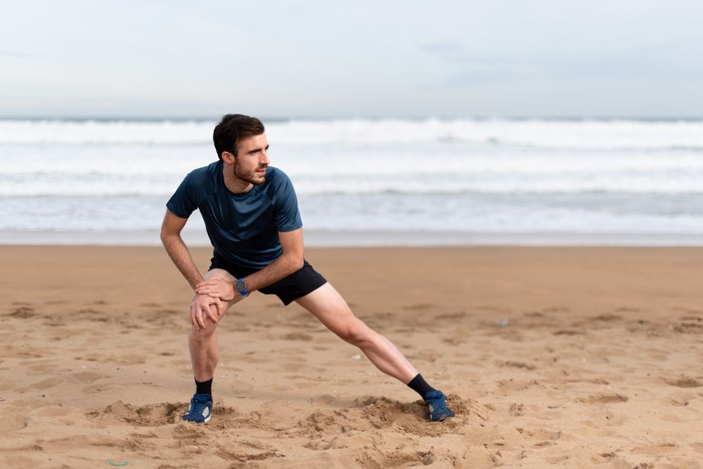 Male athlete in active wear doing exercises on legs at empty beach