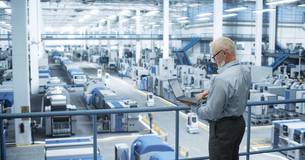 Experienced Male Engineer Standing on a Platform, Using Laptop Computer and Overlooking Production at a Modern Automated Electronics Manufacture with AI Support. Factory Executive at Work