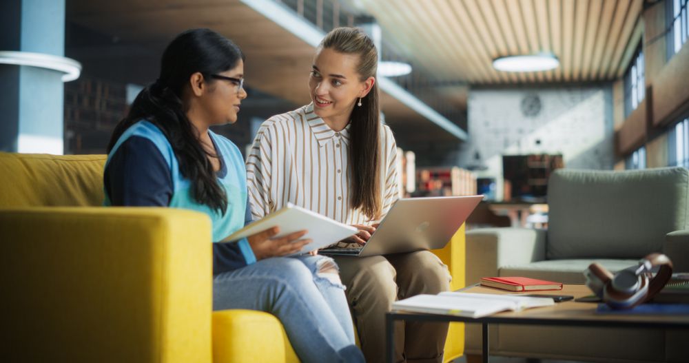 Two Diverse Friends and Classmates Working on a Collaborative Team Project in an Academic Library. Young Indian and Women Using Laptop Computer for Online Learning and Virtual Lectures