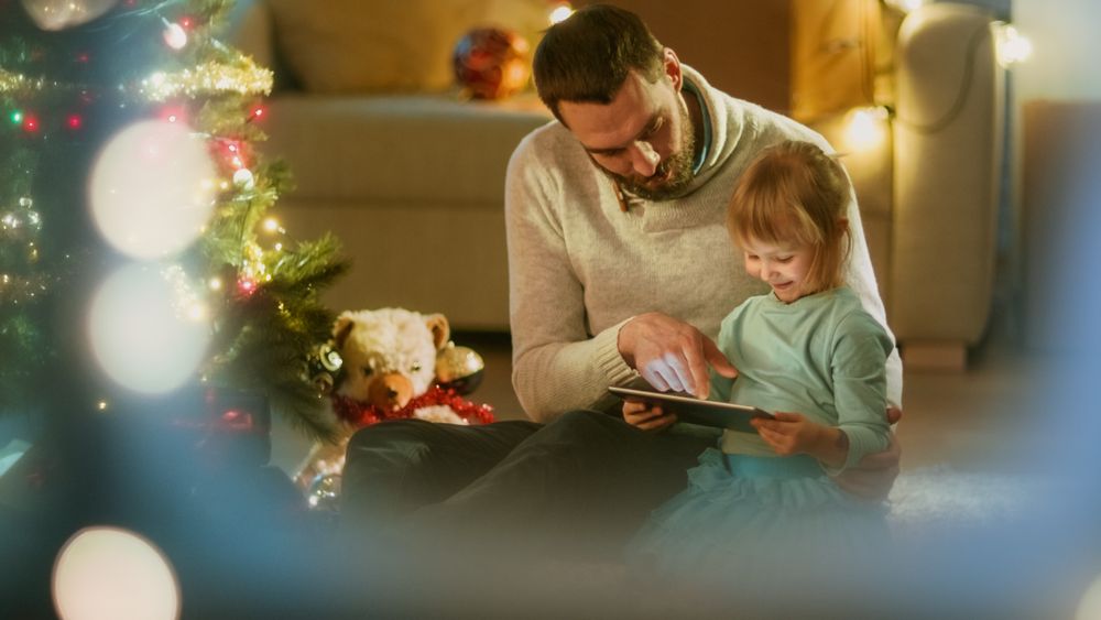Sitting Under Christmas Three with Gifts Under It Father Uses Tablet Computer with His Daughter.