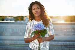 Serious Black woman holding a blue flower on a sunny rooftop