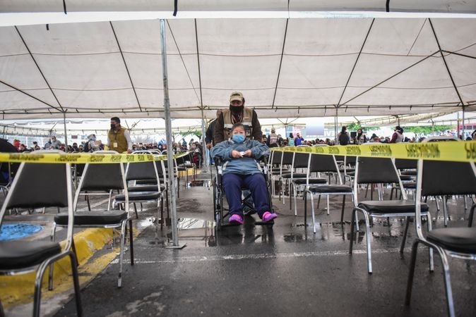 Man with facemask with an older woman on wheelchair walking between chairs