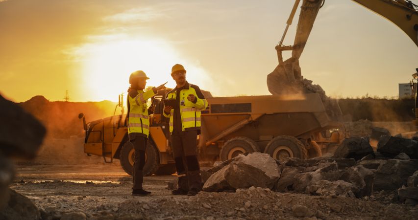 Cinematic Golden Hour Shot Of Construction Site: White Male Civil Engineer And Hispanic Female Urban Planner Talking, Using Tablet. Trucks, Excavators, Loaders Working To Build Apartment Block