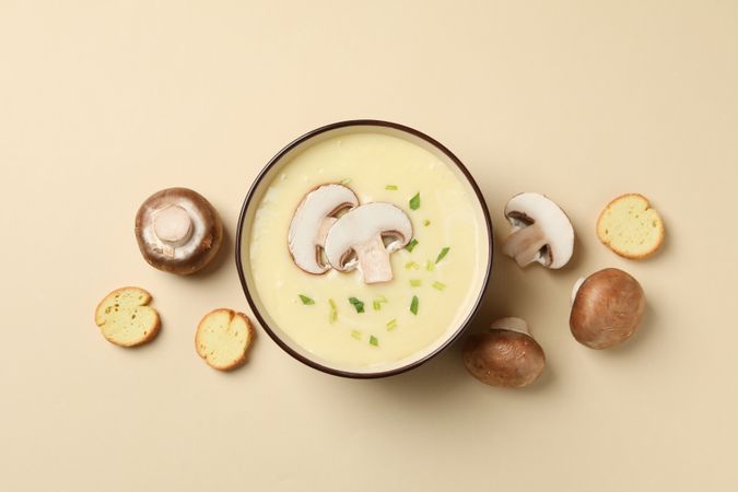 Looking down at bowl of mushrooms surrounding bowl of soup on beige background