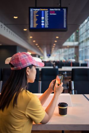 Asian woman browsing smartphone while drinking coffee at table under lighting lamp