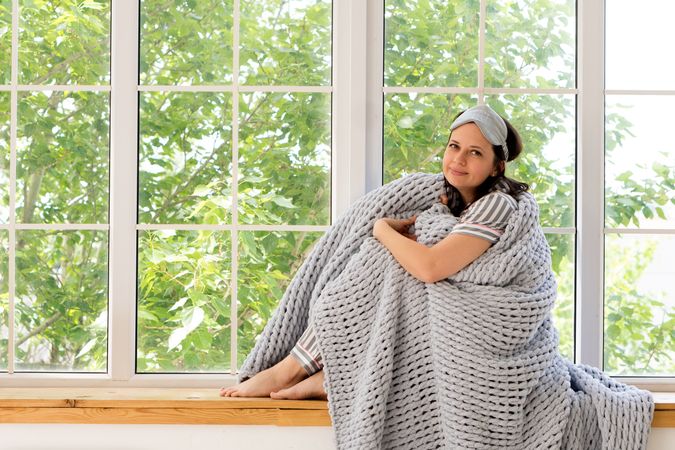 Woman smiles warmly, wrapped in chunky gray blanket by window.