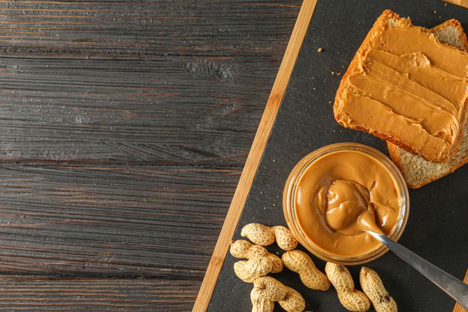 Glass jar with peanut butter and spoon, peanut, peanut butter sandwich and cutting board on wooden background, space for text and top view
