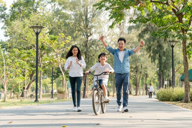 Cheerful father raises hands up in the air to support son on bike in park