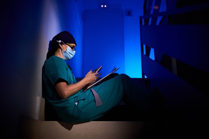 Woman in mask and scrubs sitting on stairs