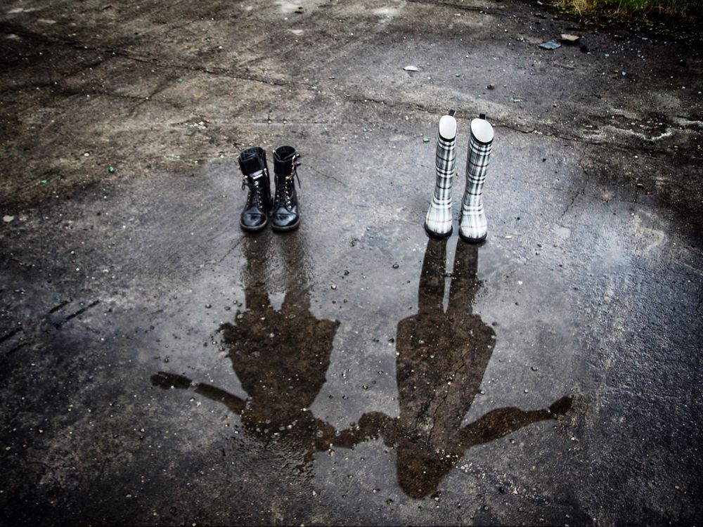 Two pair of shoes on ground with reflection of two girls on water on ground from the rain