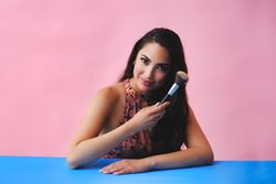 Hispanic woman with long brown hair holding large make up brush up and looking at camera