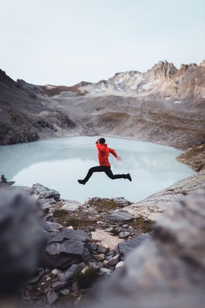 Side view of a man in red jacket jumping beside frozen lake in the mountain