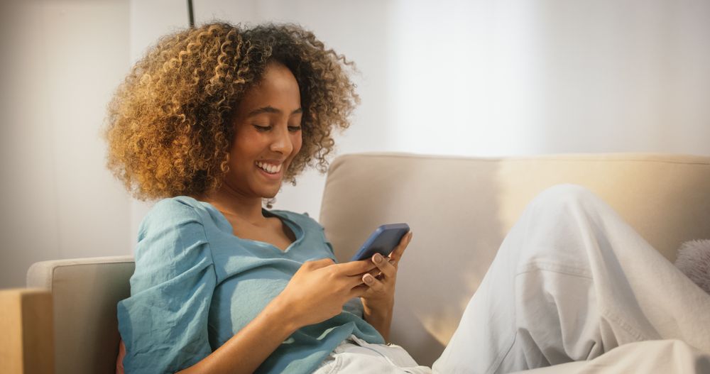 Happy Black Female Using Smartphone in Cozy Living Room at Home.