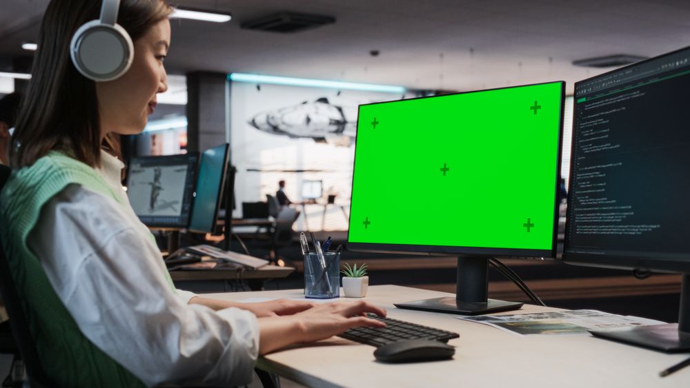Female Asian Game Programmer Coding On Desktop Computer With Green Screen Chromakey On Display In Game Development Studio Office.
