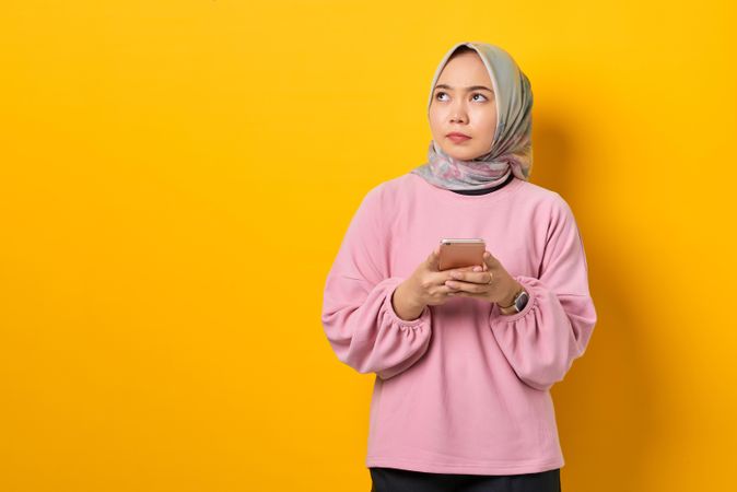 Muslim woman in headscarf holding smart phone and looking up in thought