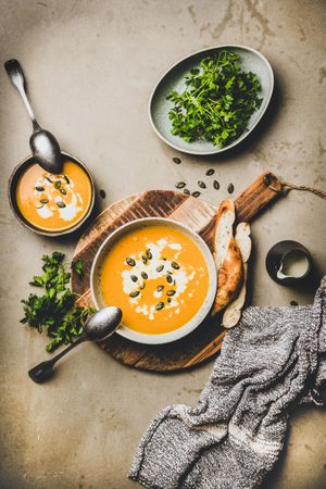 Two bowls of pumpkin soup with garnishes, bread, cream on concrete surface