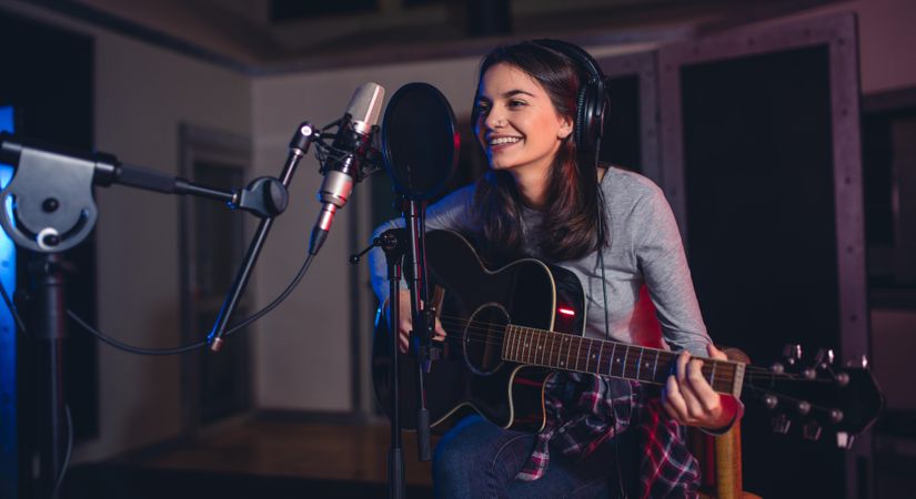 Smiling female sitting on front of microphone with guitar in recording studio