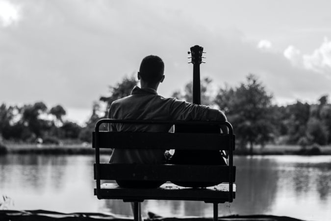Back view of man with guitar sitting on bench by shoreline in grayscale