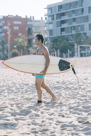 Male surfer strolling on beach with board attached to his leg with leash