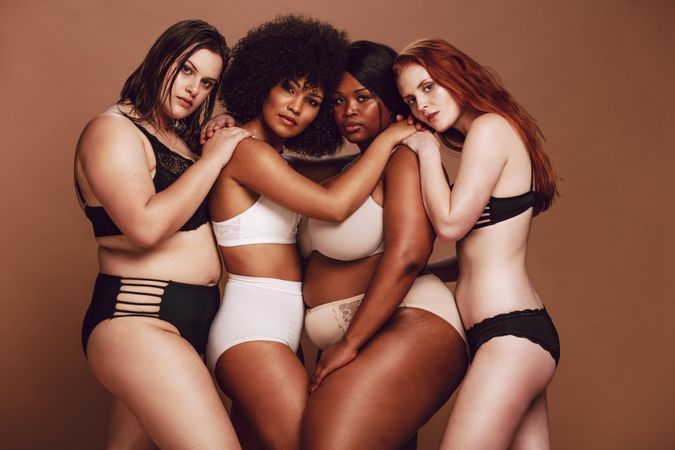 Multi-ethnic group of females with arms on each other’s shoulders posing in lingerie