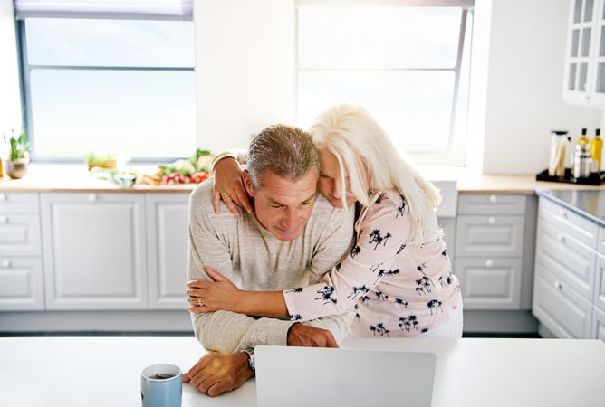 Older couple embracing in front of laptop in bright kitchen