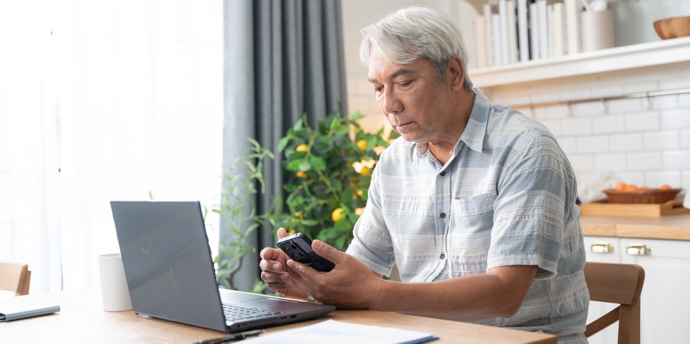 Asian mature man checking on mobile phone while working with laptop at home