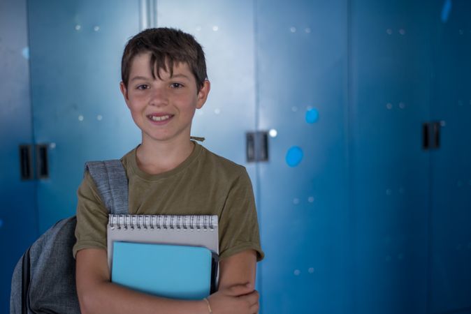 Teenager smiling while holding his notebooks in front of blue school locker