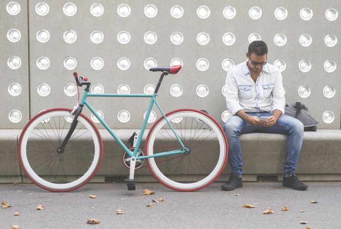 Male checking phone while sitting with bike parked in front of patterned cement wall