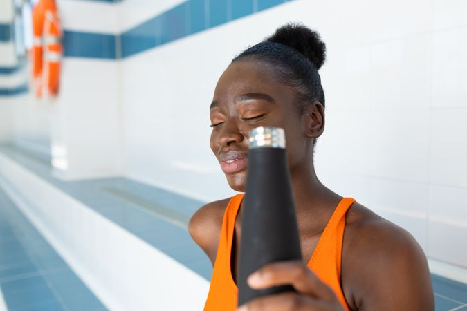 Black young woman holding her bottle of water in hand, while sitting in a bench at the indoor swimming pool