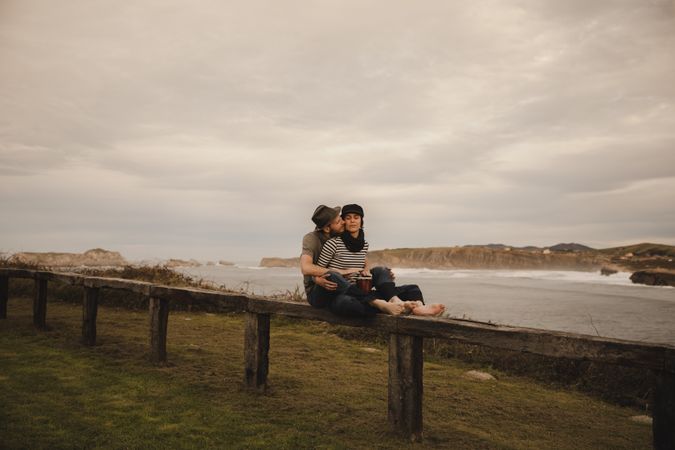 Young guy in hat near elegant woman playing on hand drum in cap on seat near coast of sea and cloudy sky