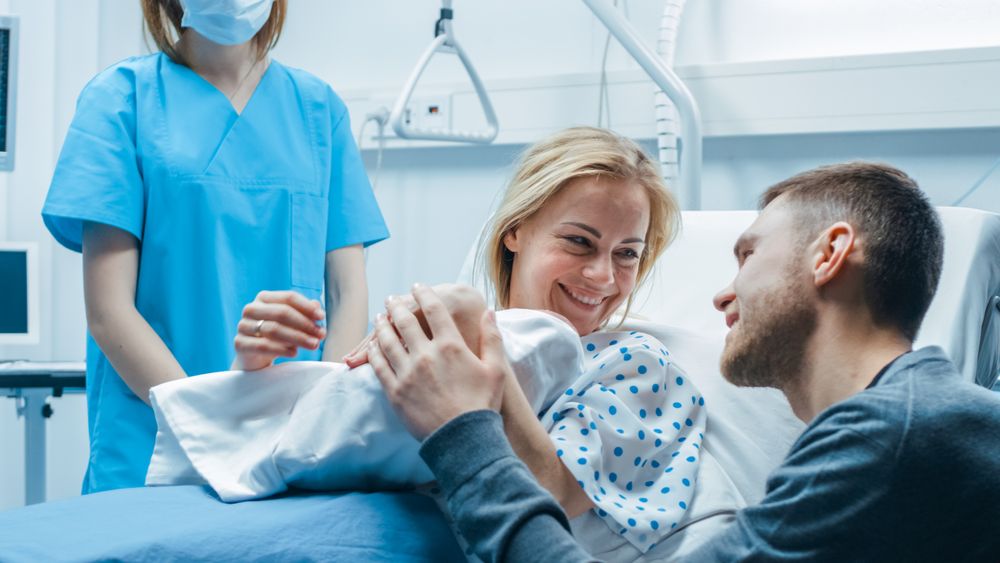 In the Hospital Midwife Gives Newborn Baby to a Mother to Hold, Supportive Father Lovingly Hugging Baby and Wife