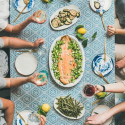 Group of people at blue patterned table with fish, and vegetable side dishes, square crop