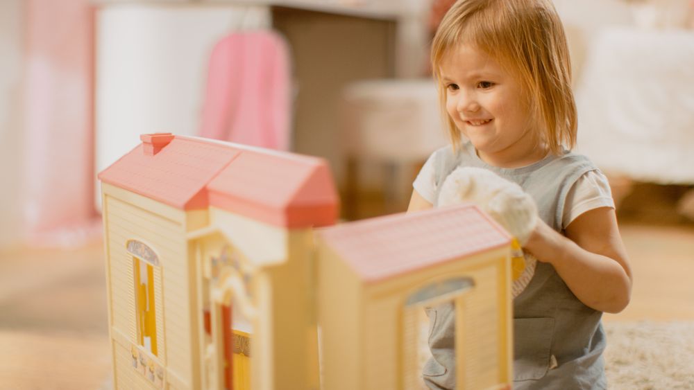 In the Children's Room: Happy Little Girl Playing with Doll House and Sitting on a Carpet.