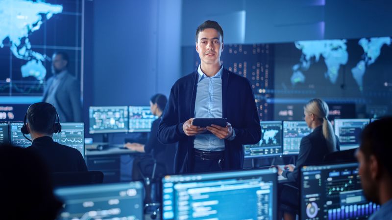 Professional Holds Tablet in High-tech Operations Center. Team Monitors Global Data on Screens. Blue-lit Environment With World Map Displays. Cutting-edge Technology for Real-time Analysis.