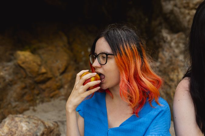 An orange-haired woman bites into an apple enjoying the outdoors