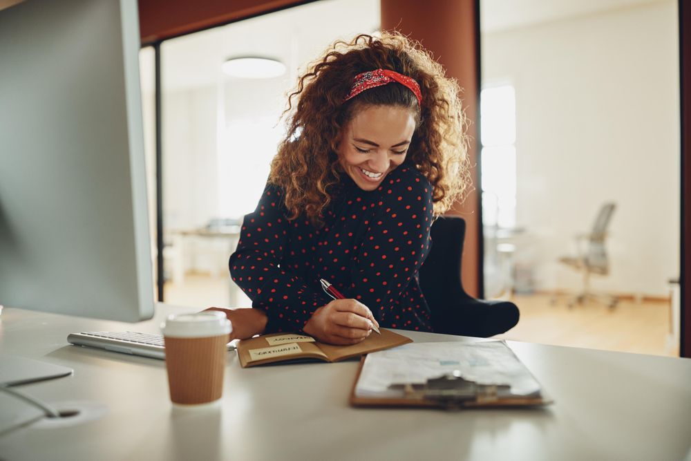 Woman with curly hair smiling while writing notes at her work computer