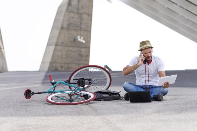 Male sitting outdoors using phone with bicycle and using laptop