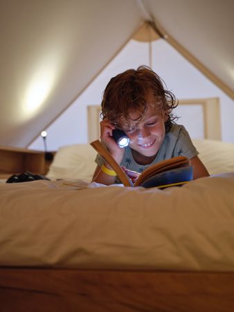 Cheerful boy reading book with flashlight