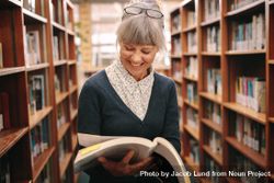 Cheerful mature woman standing in a library looking at a textbook 48aRYb