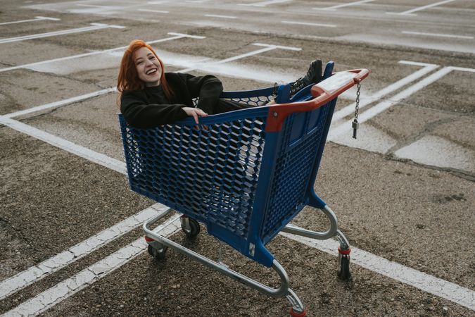 Smiling woman sitting in shopping trolley in parking lot