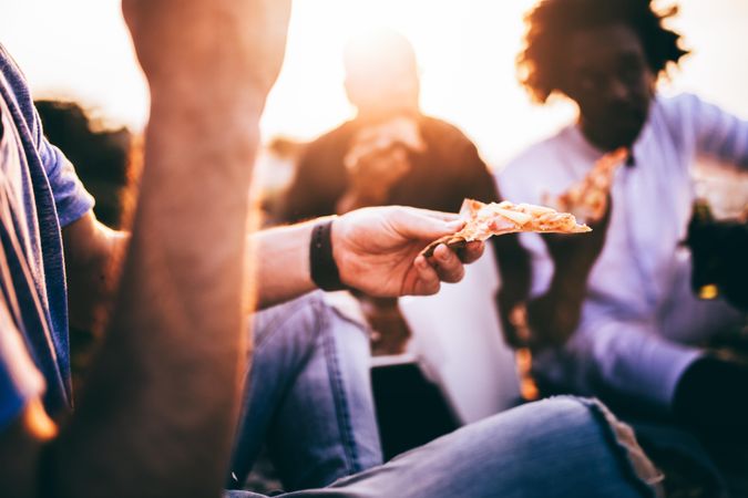 Slice of pizza is the focus among a group of friends in the foreground at sunset