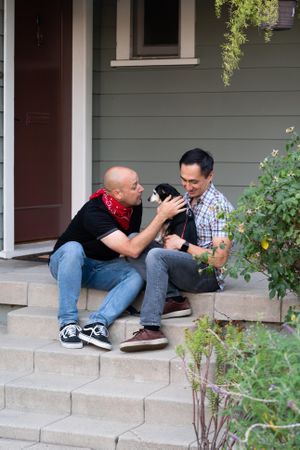 Married couple sit with their dog on front porch of their home with plants in foreground