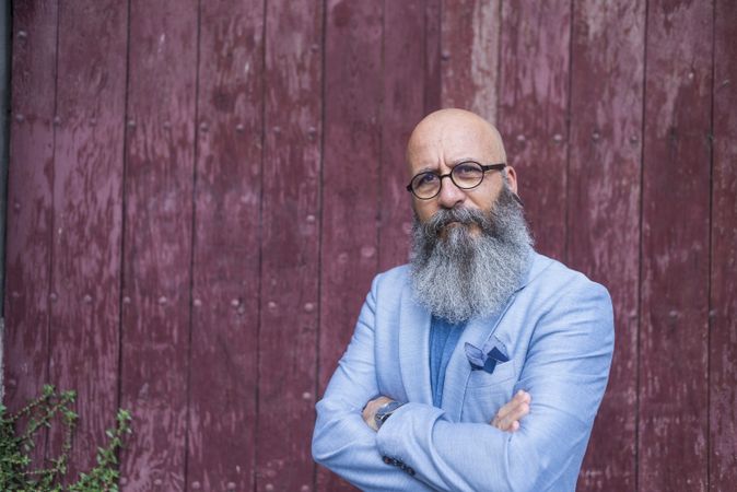 Front view of a mature trendy bearded hipster standing against a vintage red door outdoors while looking to camera