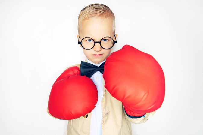 Serious blond boy wearing round glasses putting his arms out with red boxing gloves
