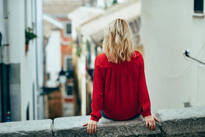 Back of blond woman sitting on stone wall with legs crossed