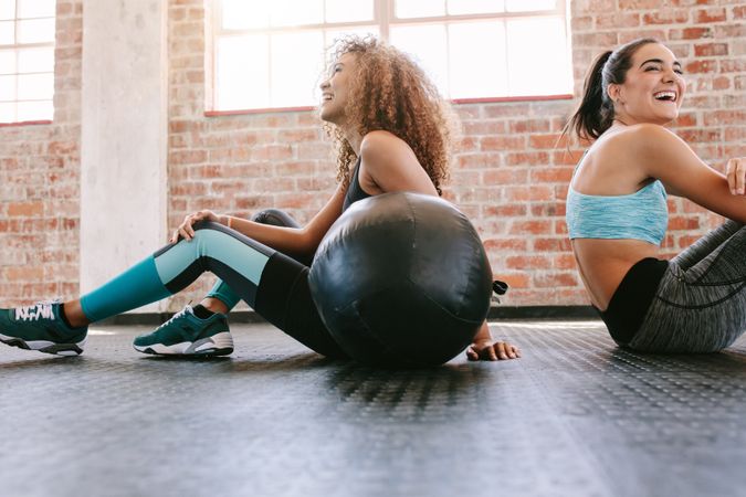 Two female friends sitting on gym floor with medicine ball and smiling